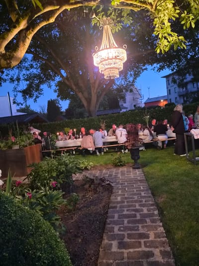 The image depicts an outdoor evening gathering at a long table set for a meal in a garden, likely in the city of Bettembourg, Luxembourg. A large, ornate chandelier hangs from a tree, illuminating the scene. Several people, appearing to be adults and some children, are seated at the table, engaged in conversation and dining. The table is laden with food, drinks, and lit candles, suggesting a celebratory or social occasion. The garden setting includes lush greenery, a brick pathway, and a hedge. In the background, buildings and trees are visible under a twilight sky. The overall atmosphere is one of pleasant conviviality.