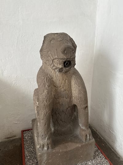 A stone sculpture, possibly depicting a stylized creature or animal, stands vertically in an indoor setting. The sculpture is made of a grayish, textured stone with intricate carvings, particularly visible on its body, which suggest scales, feathers, or fur. The head features a distinct, deep-set eye socket containing a dark, circular object, possibly metal. The creature appears to be seated or standing on a simple, robust base. Beneath part of the sculpture, a rectangular tray filled with small grey pebbles is visible, forming part of its display arrangement. The background consists of a plain, light-colored (white or off-white) wall, likely plastered, showing some texture. The lighting is even, typical of an indoor environment with artificial or diffused natural light. The overall scene suggests an exhibition or museum display, consistent with its context in Tepotzotlán, Mexico. No activity is occurring, and no text is visible in the image.