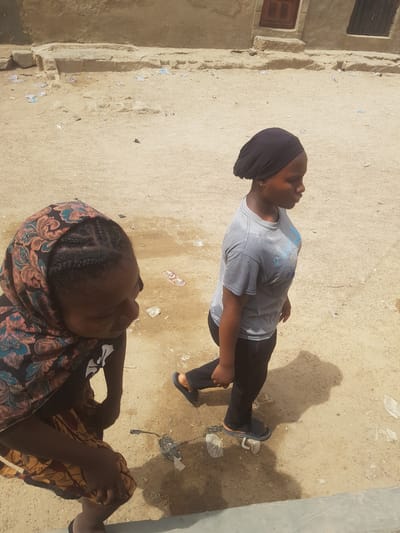 Two individuals are in an outdoor area in Dutsinma A, Nigeria. One person stands wearing a gray t-shirt, black pants, and a black head covering. The second person is seated, wearing a patterned shawl and a black shirt. The ground is sandy and shows signs of debris. A building with a small window is visible on the left side of the frame.