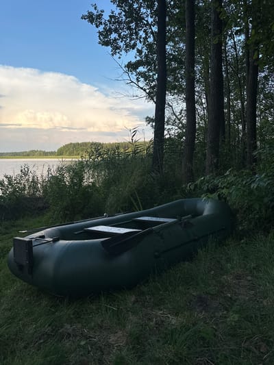 An inflatable, dark green boat rests on a grassy shore beside a body of water. The boat is positioned diagonally in the frame, with its front end facing left and its stern to the right. Oars are visible inside the boat, lying across a wooden slat. In the background, a dense line of trees forms a backdrop, with their dark silhouettes contrasting against a bright, cloudy sky. The water appears calm and reflects the muted tones of the sky and surrounding foliage. The environment suggests a natural, possibly rural, setting. The location is identified as Слабодкаўскі сельскі Савет, Belarus.