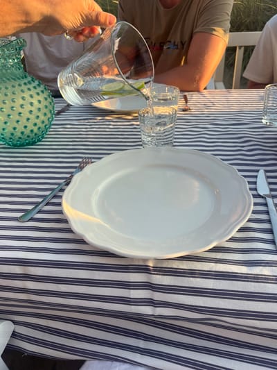 An outdoor dining table is set with a blue and white striped tablecloth over a white base. In the foreground, a large, white, fluted oval plate is positioned centrally with a silver fork to its left and a silver knife to its right. To the left, a light blue glass vase with a hobnail texture is present. A clear glass pitcher, containing water and lime slices, is being tilted by an unseen individual's hand, actively pouring water into a clear glass tumbler. Another clear glass tumbler is partially visible on the right side of the table. In the background, two individuals are seated at the table; one wears a beige t-shirt with a dark graphic, and the other's arm is visible. Behind them, white slatted chairs are discernible against a backdrop of natural sand dunes with sparse, tall grasses, illuminated by warm, low-angle sunlight. The scene is located in a coastal area, characteristic of Skagen, Denmark.