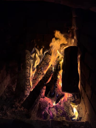 The image depicts a fire burning inside a brick fireplace. Several logs are arranged within the fireplace, with flames emanating from them. The logs range in color from dark brown to black, some glowing red and orange where the fire is most intense. Some embers appear purple and orange. The interior of the fireplace is dark. The bricks surrounding the fire are visible, creating the framework for the hearth. The image was taken in the city of Lilkovo, Bulgaria.