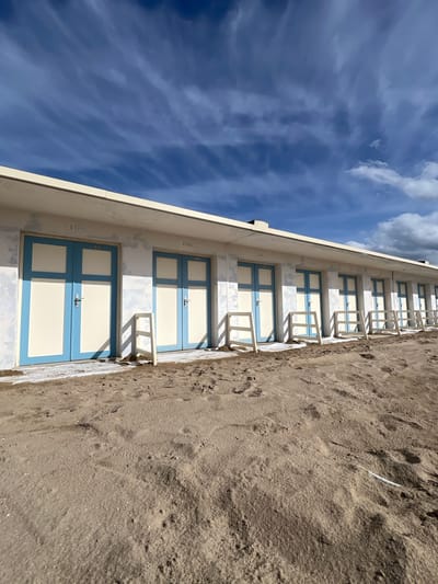 This is a medium-angle, eye-level, full shot of a row of beach cabins numbered 100, 99, 98, 97, 96, 95, 94, 93 and others less visible, located on the beach in Trouville-sur-Mer, France. The cabins are white with pale blue trim around the doors and windows. Each cabin has a single door with two panes of glass at the top. There are white railings in front of each door. The cabins are situated on a sandy beach, with footprints and some debris visible in the sand. The sky above is a deep blue with wispy white clouds. The overall impression is one of a sunny day at the seaside.