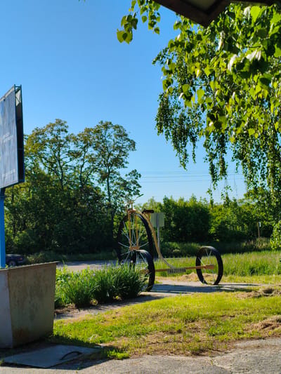The image depicts a bright, sunny outdoor scene in Velyka Bahachka, Ukraine. The central subject is a distinctive, oversized three-wheeled structure, resembling an artistic interpretation of a bicycle or tricycle. It features one very large spoked front wheel and two smaller spoked rear wheels, with a frame made of a light-colored material, possibly wood. On the rear part of this structure, a small white plate bears the black inscription "B52019BB".

To the left, a tall, blue-framed billboard is visible, with faint, illegible text on its surface. In the left foreground, a large, utilitarian, rectangular bin or container, made of weathered light-colored material, rests on the ground.

The background is dominated by lush green trees and dense bushes, creating a natural, verdant setting under a clear, bright blue sky. Faint power lines stretch across the distant sky. The top right corner of the frame is naturally adorned by hanging green leaves from a tree, likely a birch, casting a dappled light effect. The ground is covered with green grass, intermingled with drier patches, and a worn concrete or asphalt path runs through the scene.

The time of day appears to be midday or early afternoon, characterized by strong, clear sunlight. The weather is bright and clear. No human activity is present, lending a tranquil atmosphere to the scene.