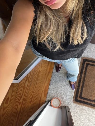 A high-angle image depicts a standing individual in an indoor entryway in Oliveira de Frades, Portugal. The subject, viewed from the mouth down, has light blonde hair, pink-toned lips, and fair skin. They are wearing a black top with lace detailing, light blue jeans, and dark red T-strap flat shoes. The left arm is raised and extends into the upper left quadrant of the frame. The floor features a light-toned wooden section with a visible grain on the left, separated by a white-framed threshold from a speckled light gray floor covering the right half of the image. A brown doormat with a dark rectangular border is positioned on the far right of the speckled floor. A small, unlit, round candle in a terracotta holder rests on the speckled floor near the base of the white frame.