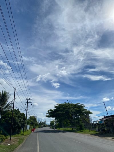 The image captures a view of a two-lane asphalt road stretching into the distance under a bright blue sky dotted with wispy white clouds. Numerous power lines crisscross the frame, suspended from utility poles on either side of the road. Dense green trees line the left side of the road, with some taller palms visible in the foreground. On the right, there are several small structures that appear to be shops or stalls, with some displaying signage. A large, mature tree with a wide canopy stands prominently in the mid-ground on the right. The road appears to be a thoroughfare in a semi-urban or rural area, possibly located in Payarangkuluh, Indonesia, given the context. There are no visible people in the foreground, but in the distance, there are a few vehicles, including motorcycles and what appear to be trucks, suggesting ongoing traffic. The sun is visible in the upper right corner, casting a bright light and creating some lens flare. The overall impression is a snapshot of a typical day on a road in a tropical or subtropical region.