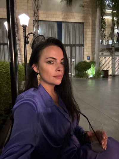 A woman with dark hair and makeup is captured in a medium shot, looking towards the camera with a direct gaze. She is wearing a deep purple, collared, long-sleeved satin shirt and a matching skirt. A delicate gold necklace is visible around her neck, and she wears a pair of gold geometric earrings with dangling feather-like accents. Her right hand rests on a clear glass containing a light-colored drink with a black straw.
The setting appears to be an outdoor plaza or courtyard in Beer Tuvia Regional Council, Israel. The background features architectural elements with cream-colored brickwork and large windows covered by gray curtains. A street lamp with an ornate glass shade illuminates the foreground. To the right of the woman, palm trees and lush green bushes are visible, with artificial lighting subtly highlighting the foliage. In the distance, a balcony with railings is visible, with people standing on it. The ground is paved with light gray concrete.