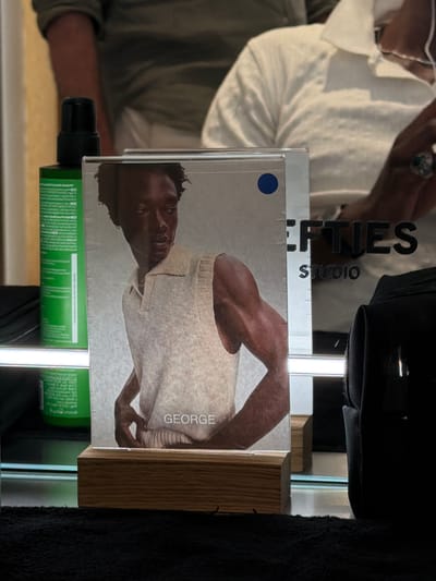 The image shows a framed photograph of a Black man wearing a sleeveless collared shirt, positioned on a wooden stand. The photograph is displayed on a reflective surface, likely a counter or shelf, in what appears to be a salon or barbershop. To the left of the photograph, a green bottle with a black pump dispenser is visible. In the background, a person wearing a white shirt and a partially visible person in a green shirt are reflected in a mirror. The text "STUDIO" and "LEFTIES" are visible on a surface behind the photograph. The scene is brightly lit, with a focus on the displayed photograph and its immediate surroundings. There is no explicit indication of the specific location within Fibracolor, Spain.