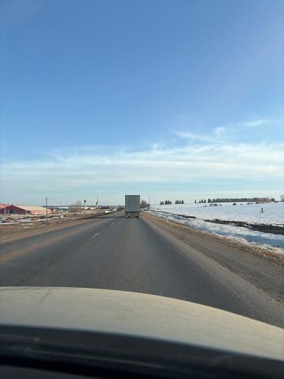 The image depicts a road, likely within or near Bol'shaya Sosnova, Russia, given the presence of buildings and infrastructure consistent with a rural Russian setting. A gray truck is visible on the road, traveling in the same direction as the camera. The road surface is dark gray asphalt, and there is a white lane marker visible. To the left of the road, there are buildings, some of which are red, and scattered patches of snow, indicating it is likely early spring. To the right of the road is a snowy field. The sky is blue with some light, wispy clouds. Part of the vehicle the image was taken from is visible on the right side, obscuring some of the view.