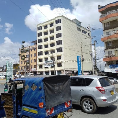 The image depicts a street scene in Ongata Rongai ward, Kenya, under a partly cloudy sky with bright blue patches and white cumulus clouds.

In the foreground, a blue three-wheeled auto-rickshaw (tuk-tuk) is positioned to the left of center, facing away from the viewer. It has a black canvas cover over its rear passenger compartment, and its rear features red and white reflective tape along with a yellow license plate displaying "KYB" (partially visible). To its immediate right, a silver Toyota RAV4 SUV is oriented towards the right side of the frame, showcasing its rear with a yellow Kenyan license plate "KDA 534Y".

The midground and background are dominated by multi-story commercial and residential buildings. A prominent, tall, light-cream colored building occupies the central background, featuring numerous evenly spaced, dark-framed windows. A sign on its ground floor identifies it as "PALM TREE HEALTHCARE - NOW OPEN." To the left of this building, a taller, yellowish-brown multi-story structure is visible, topped by an additional rooftop component. Further to the left, a vertical white sign with green text lists various medical services, including "OP HOSPITAL," "MATERNITY," "ULTRASOUND," "X-RAY," "LABORATORY," "CT-SCAN," "DIAGNOSIS," "PHARMACY," "DENTAL," and "AMBULANCE." To the right of the central cream building, another multi-story building with distinct orange-colored balconies and white accents is present. A sign on its lower level displays "MIRA STORE" and "TRINITY."

Numerous wooden utility poles supporting a complex network of overhead power and communication lines crisscross the upper portion of the image. The street surface appears paved, and additional vehicles are partially visible in the distance. Sparse green foliage is discernible near the yellowish-brown building on the left. No identifiable individuals or specific actions are visible.