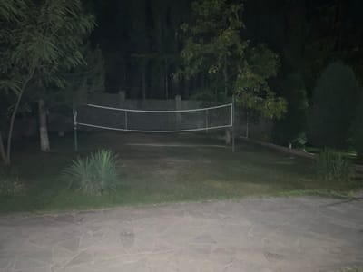 The image depicts a volleyball court at night, set within a grassy area surrounded by trees. The court has a net stretched across its width, supported by poles. A paved walkway made of irregular, light-colored stones is visible in the foreground, and the background features a fence and more trees. The lighting is dim, suggesting the presence of artificial illumination, possibly from nearby lights not visible in the frame. There are no people or animals present in the scene. The environment appears to be a park or a recreational area.