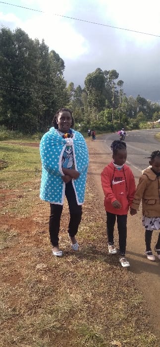 Three individuals stand on a path next to a grassy area. The person in the foreground wears a light blue wrap with white polka dots and a white trim. Behind her, a child in a red sweater stands on the path. A third person, likely a child, stands partially visible behind the second child, wearing a brown jacket. Trees and a cloudy sky appear in the background.