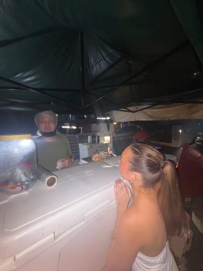 The image captures an outdoor market stall scene at night in St. Lawrence, Barbados. The photo is oriented vertically, showing a dark green canopy or umbrella structure on the left side, indicating an outdoor setup.

In the right foreground, a woman with light brown hair tied in a ponytail leans over a large white cooler. She wears a white strapless top, and her hands, adorned with rings, rest on the cooler's surface as she appears to look down into it. The cooler bears the visible text "TAHOE TROUT" and "ARCTIC SERIES".

Behind the cooler stands a man, likely a vendor, wearing a light-colored head covering or turban and a green shirt. He has a beard and gazes towards the camera with a slight smile, suggesting a friendly demeanor. To his right, a clear plastic dispenser filled with colorful small items, possibly candy or beads, sits on or near the cooler.

The background is dark and blurred, with various indistinct lights suggesting other market stalls, buildings, or streetlights in the evening setting. The scene conveys a casual, interactive atmosphere at a lively outdoor market.