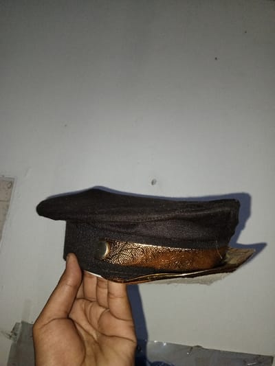 An individual's hand holds a dark-colored peaked cap against a light grey wall in Na Sang, Laos. The cap features a wide, gold-colored band with an intricate embossed pattern, secured by a circular, silver-toned button on the wearer's left side. The cap's crown and brim are made of a dark fabric, possibly black or dark navy. The hand, exhibiting a medium skin tone, grips the cap from below, with the thumb visible on the left side near the button and fingers supporting the brim. A soft shadow of the cap and hand is cast upon the plain, textured wall in the background.