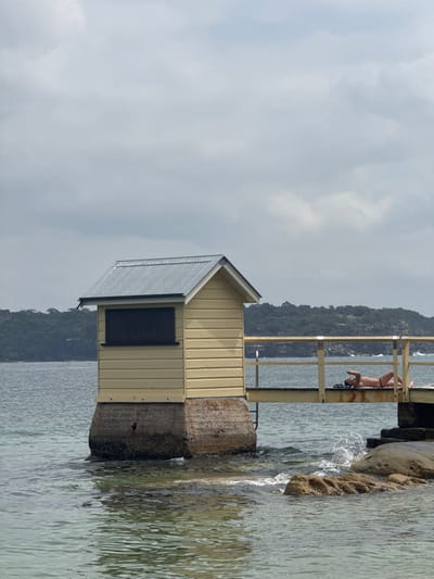 This image depicts a coastal scene in Watsons Bay, Australia, under a uniformly overcast sky. A single-story building, clad in light yellow horizontal timber, features a dark grey corrugated metal gabled roof and a dark, shuttered window on its visible facade. This building rests on a robust concrete plinth situated in the water. A light yellow wooden pier, equipped with railings, extends from the right side of the frame, connecting to the building's foundation. A sole individual, an adult male, is positioned supine on the pier's wooden deck, clad in dark swimming briefs. His right arm is flexed with the hand near his head, and dark footwear or accessories are present beside him. The surrounding water is relatively calm, displaying ripples and small waves breaking against the pier's support and exposed natural rock formations in the foreground. A distant, heavily vegetated landmass forms the background across the body of water.