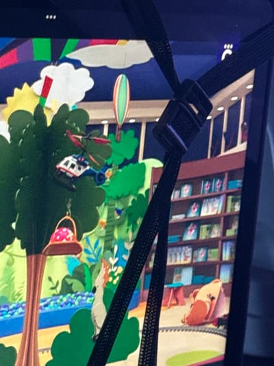 A close-up photograph captures a section of a screen or display showing a vibrant, multi-layered illustrated scene, partially obscured by a black strap with a plastic buckle crossing diagonally from top-left to bottom-right. The strap originates from a dark blue surface on the left, possibly the device's bezel, where a white symbol resembling ":C" and three vertical dots are visible.
The illustrated scene depicts a whimsical, natural environment combined with interior elements. In the upper right, a cartoonish, light brown animal, possibly a chipmunk or squirrel, wearing a hat, peeks from behind a structure. Behind it, a brown bookshelf or display cabinet holds numerous small, colorful items, suggesting books or pictures, arranged in rows.
The central and lower portions of the illustration feature lush green foliage, including a prominent tree. A small white and blue helicopter with red propellers hovers near the tree. A red and white spotted object, possibly a basket or mushroom, hangs from a branch. To the right, a long, slender gray animal with an orange back, resembling a ferret or weasel, lies horizontally. A blue, textured vertical band, possibly representing a stream or river, runs through the scene.
Other elements include an oval-shaped, striped object (green, orange, white) floating in the air, a white cloud-like shape with a red and green stick-like attachment, and a section with colorful vertical stripes (purple, green, red, yellow) on the far left. The art style appears to be paper cut-out or digital illustration, creating a playful and detailed landscape. No specific activity is clearly depicted beyond the presence of these elements in their setting.