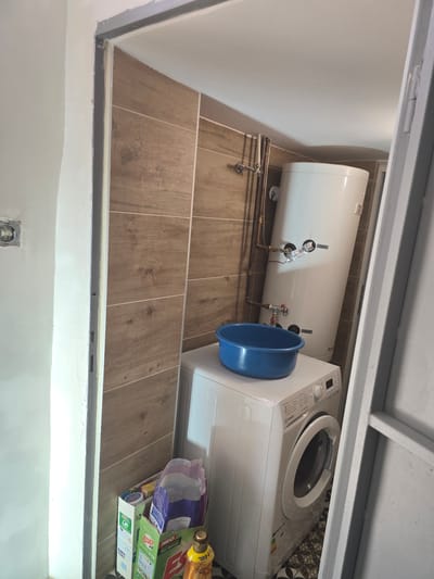 A storage closet in Gorno Aleksandrovo, Bulgaria, contains a horizontal hot water heater, a washing machine, a blue plastic basin, and assorted laundry supplies. The space is enclosed with light-colored walls and a brown tiled floor. Visible pipes and valves connect to the hot water heater. The washing machine is white.