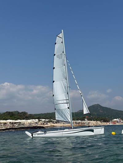 A white catamaran sailboat with a tall mast and white sail is anchored in the clear blue water off the coast of Castiadas, Italy. The boat is positioned in the foreground, with the beach and lush green hills in the background. The beach is lined with white umbrellas and lounge chairs, suggesting a popular tourist destination. A few people are visible on the beach, relaxing under the umbrellas and enjoying the sunny day. The water is calm, with gentle waves lapping against the boat. The sky is a clear blue, with a few fluffy white clouds scattered across the horizon. The overall scene is one of a peaceful and idyllic beach vacation.