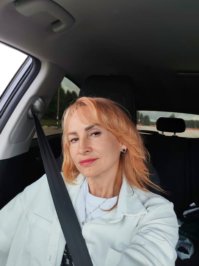 A medium shot captures a Caucasian woman with auburn hair and fair skin, wearing a white jacket, a white t-shirt, and a silver necklace, seated in the passenger seat of a car. Her seatbelt is fastened. She is looking directly at the camera with a neutral expression. The car's interior is visible, including a black headrest and interior trim. Outside the car, trees and a road are discernible. The image appears to be taken on a cloudy day. The location is not specified in the image.