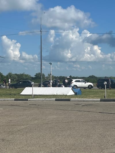 The image shows a street scene with several cars parked along a roadside. People are visible near some of the vehicles, suggesting some kind of gathering or event. The sky is blue with scattered white clouds, indicating fair weather. The time of day appears to be daytime, likely afternoon, given the lighting conditions.
Several cars are present, including a white SUV and a dark-colored sedan. There's also a white structure with a corrugated surface, possibly a temporary barrier or display. Power lines are visible stretching across the sky, supported by poles, and a street light is also present. The ground is paved with asphalt, and a grassy area borders the road.
The presence of people gathered near the cars suggests an informal event, perhaps a car show, a community gathering, or simply people socializing. The overall atmosphere appears casual. No specific text is legible in the image. The location is identified as Temryuk, Russia.