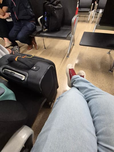 The image shows a partial view inside what appears to be an airport or waiting area, likely in Rionegro, Colombia, due to the prompt context. A person wearing light blue jeans and red canvas shoes is sitting with their luggage next to them. Another person is seen lying down in the seating area, dressed in dark clothing and brown shoes. The floor is a neutral beige color, and there are airport chairs with black seats and metal frames. A water bottle can be seen under one of the chairs. A black suitcase with a visible handle is positioned on the floor. The scene conveys a sense of waiting and travel.