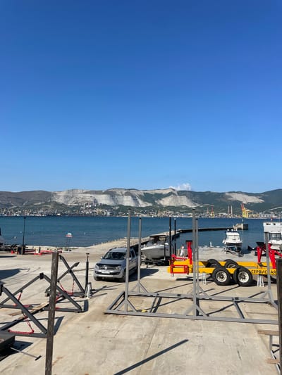 An outdoor scene on a sunny day shows a body of water with boats docked and a distant city or industrial area on the other side. In the foreground, a concrete area has several metal structures, a silver SUV, and a yellow trailer with four wheels. The sky is clear blue. The scene appears to be a port or marina in Maiorovskii, Russia. There are no visible people in the foreground.