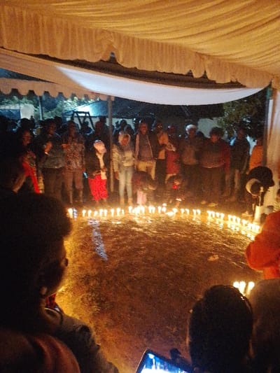 A nighttime gathering is depicted under a draped white tent structure in Marenyo-Shianda ward, Kenya. Approximately twenty individuals are present, forming an arc around a large, circular arrangement of numerous lit candles placed directly on the dark, possibly grassy or earthen ground. In the immediate foreground, the backs of two individuals' heads are visible, with one holding a smartphone displaying an illuminated screen. In the central midground, one person is observed kneeling or bending near the perimeter of the candle circle. The background, beyond the standing group, reveals faint outlines of outdoor play equipment, consistent with a swing set, against the dark environment. The scene is illuminated primarily by the lit candles and ambient overhead lighting from within the tent.