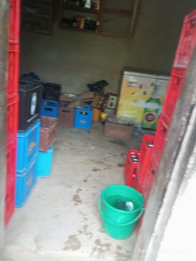 The image depicts the interior of a storage room, possibly a shop or a stockroom, in Dokan Tofa, Nigeria. The room has concrete floors and plain walls. In the foreground, on the left, are stacked blue plastic crates, some filled with what appear to be bottles or cans. A black rectangular container, possibly a cooler or a storage box, sits atop some of these crates. To the right, a bright green plastic bucket filled with water is visible. In the background, there are shelves mounted on the wall, holding various items, likely beverages or other merchandise. A large, yellow and white refrigerator or display cooler with an image of a beer bottle is situated against the back wall. There are also several brown and blue plastic crates stacked in the middle ground, some appearing empty and others possibly containing goods. Cardboard boxes are also present, suggesting inventory or packaging. The overall impression is one of a functional, perhaps slightly disorganized, storage space for retail goods. The lighting is somewhat dim, and the image appears to have been taken with a mobile phone, indicated by the timestamp "06/16/2026 15:57" in the lower right corner. There are no people visible in the image.