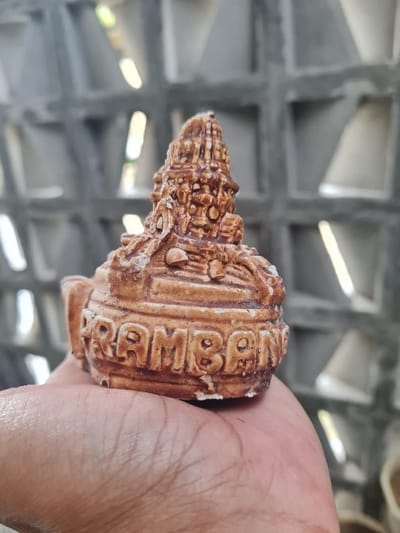 A hand holds a brown, cone-shaped souvenir with detailed relief carvings and a textured surface. The object features the word "RAMBONG" vertically inscribed in block letters along its side. The background shows a gray concrete wall with square openings that allow light to pass through. The lighting highlights the contours and details of the souvenir and the texture of the wall.