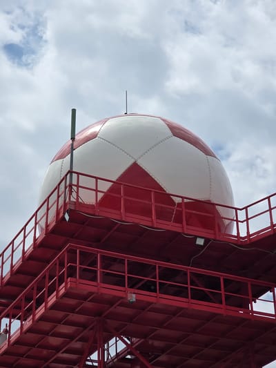 The image shows a close-up, low-angle view of a radar dome, which is spherical and painted with a red and white checkerboard pattern. The dome is mounted on a bright red metal structure with multiple platforms and railings, resembling a tower. Various antennae and other equipment are attached to the dome and the structure. The background consists of a cloudy sky. This appears to be a radar station located in or near Ignatievo, Bulgaria, which is an area known for its meteorological radar installations.