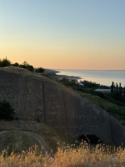 This image depicts a scenic view from atop a stone fortification, likely a historical landmark, overlooking a body of water. The sun is setting, casting a warm, soft glow across the sky, transitioning from a clear pale blue at the top to a soft orange and yellow near the horizon.
The dominant feature in the foreground is the weathered, grey stone wall of the fortification. Its rough-hewn blocks are clearly visible, suggesting age and historical significance. A dry, grassy embankment slopes down from the wall, dotted with sparse vegetation and a few small purple wildflowers. A winding dirt path is visible on this embankment.
Beyond the fortification, a calm body of water stretches towards the horizon. Along its edge, a strip of land is visible with scattered trees and the rooftops of a few buildings, suggesting a coastal settlement or town. The water's surface reflects the soft hues of the sunset.
The overall mood is peaceful and contemplative, with the natural beauty of the sunset and the serene waters. The time of day is clearly sunset, indicated by the warm light and the gradient in the sky. The location appears to be coastal, with the vast expanse of water and the historical structure suggesting a place with a rich past. No people or animals are visible in the image. There is no visible text.