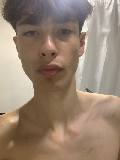 A close-up, front-facing, waist-up, selfie-style photograph depicts a young male, likely a teenager, against a backdrop of white tiled walls on the left and a white shower curtain on the right. The subject is shirtless, with his skin appearing slightly shiny, possibly due to water or sweat. His hair is dark and styled in a slightly tousled manner. His eyes are open and looking directly at the camera. The lighting in the image is somewhat harsh, creating highlights on his skin. The image does not contain any discernible elements that would suggest a specific location within the city of Glodeni, Moldova.