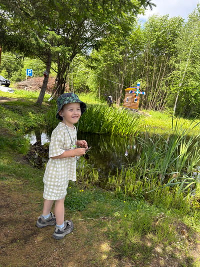 A young boy wearing a checkered outfit and a bucket hat stands near a body of water in an outdoor setting. The boy is smiling and looking to his right. In the background, there is a small wooden windmill decoration and dense green foliage. A blue parking sign is visible on the left side of the image. The ground is covered in grass and dirt. The scene appears to be a park or natural area.