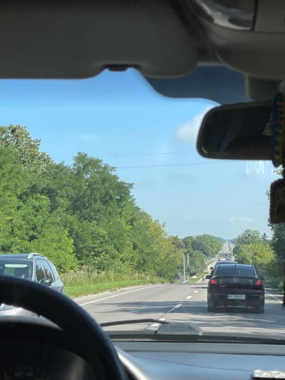 This image, taken from the perspective of someone driving a car, captures a highway scene in Sukhodoly, Ukraine. The sky is a clear blue with a few wispy clouds. Dense green trees line both sides of the road. In the foreground, the steering wheel of the car is visible. On the road ahead, several cars are driving in the same direction. A black Mazda 3, with license plate BC 3966 PI, is in the middle lane, slightly ahead of the viewpoint. Other vehicles are further down the road, receding into the distance. The road is paved and has lane markings. Streetlights are visible along the right side of the road. The overall scene depicts a typical day on a Ukrainian highway.