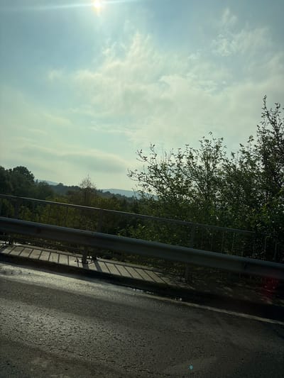 The image captures a roadside view, likely from a bridge, with a metal guardrail in the foreground. Beyond the guardrail, a landscape of rolling hills covered in dense green trees stretches into the distance under a bright, slightly hazy sky with scattered clouds. The sun is visible as a bright glare in the upper left corner. The road surface in the foreground appears wet and reflective, suggesting recent rain or dew. There are no people or animals visible in the frame. The overall impression is a natural, somewhat overcast day in a rural or semi-rural setting. The location is identified as being in the city of Ivanski, Bulgaria.