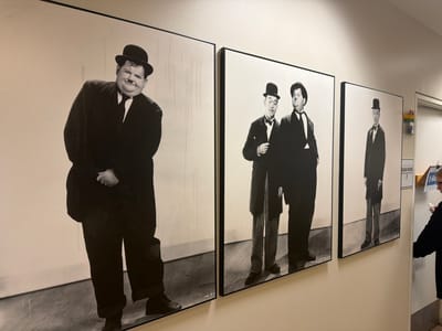 The image depicts a hallway with three framed black and white photographs on the wall. The photographs feature the comedy duo Laurel and Hardy. The first photograph, on the left, shows a close-up of Oliver Hardy from the chest up, wearing a bowler hat, suit, and tie. The second photograph shows Stan Laurel and Oliver Hardy standing side-by-side, both wearing bowler hats and suits. The third photograph, on the right, shows Stan Laurel standing alone, also in a bowler hat and suit. The hallway also has a door labeled "EXAM ROOM 2" and a sign that reads "X-RAY." A person wearing glasses and a black jacket is standing near the door, holding a white object. The scene appears to be located in a medical facility or office building, possibly in Burbank, California, given the context of the photographs.