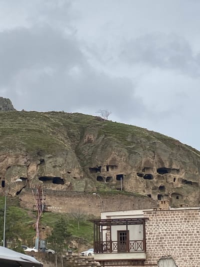 A rock formation riddled with cave dwellings rises in the city of Konya, Turkey. A building is connected to the rock face, featuring a white facade and dark wood accents. A single leafless tree is visible on the rock's peak. The sky is overcast. Modern electrical infrastructure is in the foreground. A vehicle is partially visible on the right.