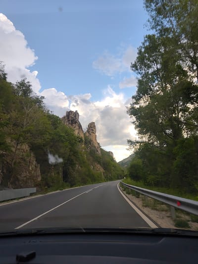 The image depicts a view from inside a vehicle driving on a paved road. The road curves slightly to the right, with a metal guardrail visible on the right side. A rock formation is visible in the background. The sky is visible above, featuring clouds. The image was taken on the road to Gospodintsi, Bulgaria.