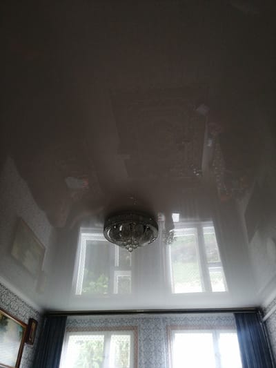 The image shows an indoor scene, likely a room in a home, with a focus on the ceiling and a portion of the walls and windows. The ceiling is a glossy, reflective surface, possibly a stretch ceiling, which mirrors the room's contents and the light sources.
Prominently featured on the ceiling is a decorative chandelier, intricately designed with multiple light sources and crystal-like elements. Its reflection is visible on the polished surface above.
Below the ceiling, several windows are visible. They are covered with sheer curtains that have a dotted pattern. Through the windows, greenery and what appears to be an outdoor landscape can be seen, suggesting daytime. The light filtering through the windows indicates it's daytime, though the overall lighting in the room is somewhat dim, possibly due to overcast weather or the time of day.
The walls appear to be wallpapered with a patterned design. A framed picture or painting is hung on the wall to the left, partially visible. Dark blue curtains are drawn to the sides of some windows.
The scene is static, with no people or explicit activity taking place. The overall impression is one of a residential interior, possibly a living room or dining area. The environment seems tidy and furnished. There is no visible text in the image. The location context of Dubrovka, Russia, doesn't offer any specific visual cues within the image itself.
