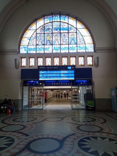 The image depicts the interior of a train station in Eisenach, Germany. The scene is dominated by a large, arched stained-glass window featuring the text "70 JAHRE Eisenacher Automobilbau" which translates to "70 Years of Eisenach Automobile Construction." Below the stained glass are rows of smaller, decorative stained-glass panels and a digital departure board displaying train information.

In the foreground, the floor is decorated with an intricate mosaic pattern of geometric shapes and circular designs in shades of red, gray, and black. To the left, two individuals are seated on a bench, with one holding a shopping bag. In the center of the image, glass-paneled doorways lead into a brightly lit hallway where several people are walking with luggage. To the right, a green ATM machine with the label "cash zone" is visible. The overall environment suggests a public transit hub.