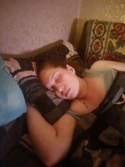 A woman is sleeping on a bed, covered by a blanket and several pillows. She is lying on her side, with her head resting on a pillow. Her eyes are closed, and her mouth is slightly open. Her hair is dark brown and is spread out around her head. She is wearing a light-colored top. The background appears to be a bedroom, with a patterned wallpaper and a decorative pillow on a sofa. The scene is dimly lit, suggesting it is nighttime or the room is dark. This image was taken in the city of Voskresenskiy, Russia.