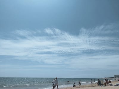 This is a wide shot of a beach in Rota, Spain, likely on a sunny day with a partly cloudy sky. The sea is visible in the middle ground, with gentle waves approaching the shore. A sandy beach occupies the foreground and extends into the background. Several people are present on the beach. In the foreground, two individuals are walking towards the water, one wearing a white shirt. To their right, a child is playing in the sand, holding a yellow object. Further to the right, a group of people are sitting on chairs, one person is wearing a black and white shirt. In the water, a few individuals are swimming or wading. In the background, buildings are visible along the coastline, suggesting a developed area. The overall atmosphere appears relaxed and leisurely, with people enjoying a day at the beach.