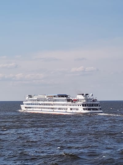 A large, white, multi-decked cruise ship sails on dark, choppy water under a bright blue sky with scattered clouds. The ship has numerous windows and balconies on each deck. A red and blue stripe runs along the waterline. The scene is set in the city of Veregonets, Russia.