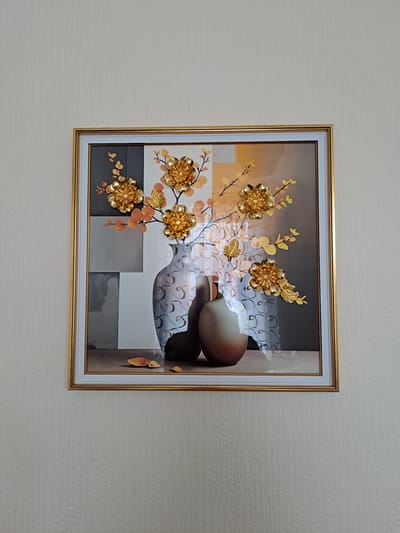 A framed picture hangs on a textured wall. The picture depicts a still life of two vases, one tall and slender with a swirling pattern, the other shorter and rounded with a gradient of brown and orange. Golden flowers with intricate petal designs and golden leaves sprout from branches emerging from the tall vase. The background of the picture features a geometric pattern of squares in shades of gray, white, and orange. The frame is golden and slightly beveled. The wall is a pale, neutral color with a subtle vertical texture.