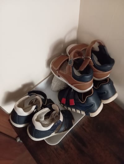 A pile of children's shoes is stacked in a corner of a room. The shoes are brown and navy blue. Some are sandals, and others are more like closed shoes. The shoes are arranged on a white surface, possibly a tray or shelf, which sits on a dark wooden surface, likely a table or dresser. The background is a plain, light-colored wall that meets another wall at a right angle. The lighting suggests an indoor setting, with soft shadows cast by the shoes and the corner of the room. There is no indication of the city of Baden, Austria, in the image.