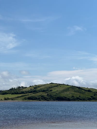 The image shows a scenic view of a lake with a green, rolling hill in the background. The lake occupies the bottom half of the frame, with its surface rippled by a gentle breeze. The water is a deep blue, transitioning to a lighter, almost reddish hue near the shore, suggesting shallow depths or a sandy bottom. The shoreline is a narrow strip of pebbles and stones.
The hill in the background is covered in lush green grass and scattered with clusters of trees and shrubs. The vegetation creates a textured pattern on the undulating slopes. A few small, white specks, likely sheep, are visible grazing on a lighter green patch on the left side of the hill. The top of the hill is dotted with small trees.
The sky above is a clear blue, with wispy white clouds scattered across it. Some of the clouds are larger and more substantial, particularly towards the horizon above the hill, where they appear to be piling up. The lighting suggests it is daytime, with the sun likely positioned out of frame, casting a soft light on the landscape. The overall impression is one of natural beauty and tranquility in a rural setting, consistent with locations in Tullycullion, Ireland.