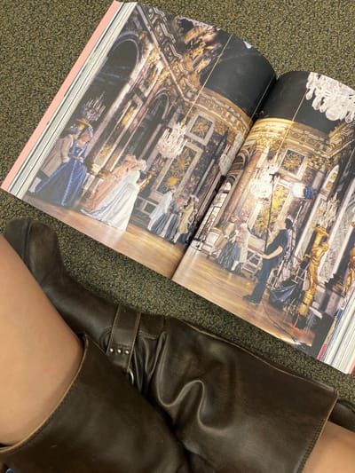 A top-down, slightly diagonal image captures an open, large-format book resting on a dark green, mottled carpet. Adjacent to the book is the lower right leg of an individual, clad in a dark brown leather boot. The book displays a two-page spread depicting an ornate interior scene, likely a historical film set or photoshoot.
On the left page, multiple figures in elaborate 18th-century style attire, including gowns and powdered wigs, are visible against a backdrop of gilded walls, dark marble accents, and crystal chandeliers. A central female figure in a light-colored gown appears to be interacting with another costumed individual.
The right page continues the interior scene, also featuring costumed figures. Crucially, a modern film crew member, dressed in a dark t-shirt, jeans, and a baseball cap, is visible operating a tall light stand equipped with a reflector or diffusion panel. Other production equipment, possibly scaffolding, is present near the wall.
The dark brown boot covers the majority of the visible leg, extending upwards from a thick sole. It features a prominent strap with a metal ring and several small metal studs around the ankle or lower calf area. The skin of the upper thigh is partially visible above the boot line. The carpet upon which the book and leg rest is dark green with subtle, irregular lighter flecks, characteristic of commercial indoor flooring.
Based solely on the visual content, it is impossible to determine a specific location within the city of El Paso, United States.
