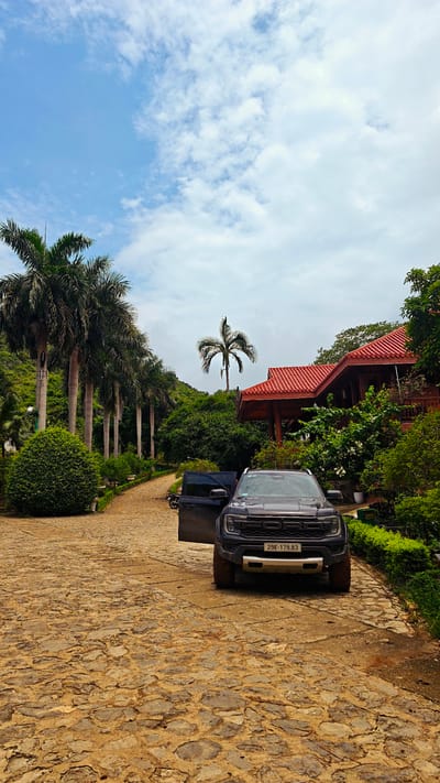 This image depicts a dark blue Ford Ranger pickup truck parked on a cobblestone driveway in front of a wooden building with a red tiled roof. The truck has its driver's side door open and its license plate, which reads "29K-179.83", is visible. The building is surrounded by lush greenery, including tall palm trees lining the left side of the driveway and a variety of other plants and trees. In the background, a hilly, forested landscape is visible under a partly cloudy blue sky. The scene appears to be a resort or a large private property, likely located in a tropical or subtropical region. While the exact location within Tam Thanh Ward, Vietnam, is not specified, the presence of palm trees and the architectural style of the building are consistent with Southeast Asian environments. No people are present in the image.
