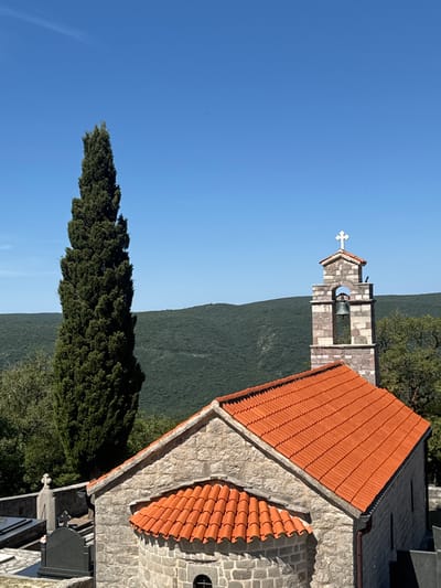 The image depicts a stone church with an orange tiled roof and a bell tower topped with a cross, set against a backdrop of rolling green hills under a clear blue sky. In the foreground, to the left of the church, are gravestones, indicating a cemetery. A tall, slender cypress tree stands to the left of the bell tower. The church appears to be situated in a rural or semi-rural setting, likely within the village of Bratešići, Montenegro, given the context provided. The scene is peaceful and serene, bathed in natural daylight. No people or animals are visible in the image.