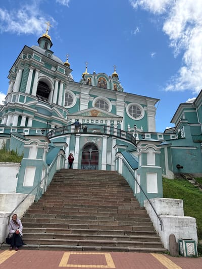 An upward-angle, eye-level, full exterior shot captures the grand facade of the Smolensk Cathedral in Demenshchina, Russia. The cathedral, painted a striking teal with white trim, features ornate architectural details, including columns, arches, and a prominent dome topped with a golden cross. A wide stone staircase leads up to the entrance, with a woman in a light-colored jacket and scarf sitting on the steps to the left, holding a red cup. Two figures are visible on the balcony above the entrance. The sky above is a vibrant blue with scattered white clouds, and lush green grass covers the embankment to the right of the stairs. The foreground is paved with red brick and yellow lines.